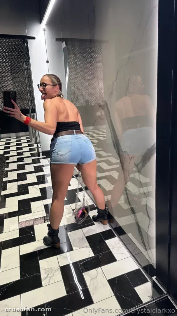Crystal Clark in a mirror selfie, wearing a black top and denim shorts in a tiled room.