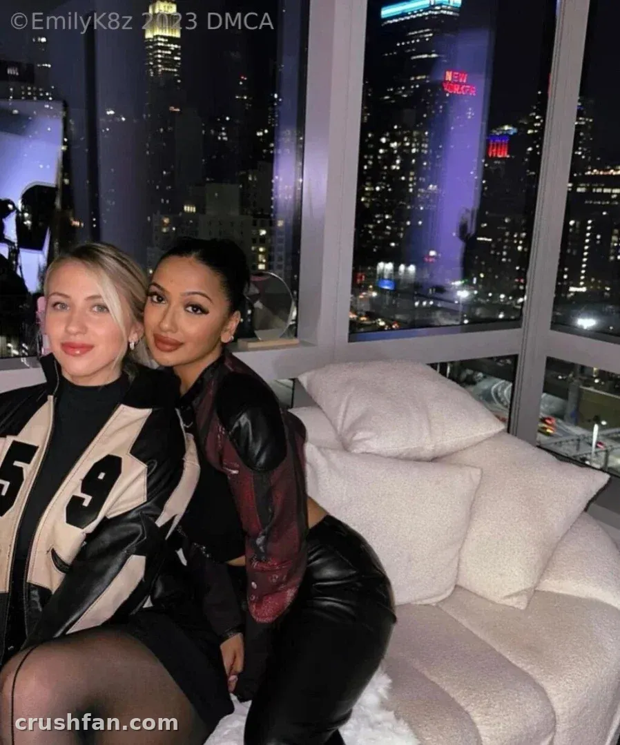 Emma K in a varsity jacket beside a friend on a sofa with a night city skyline behind them