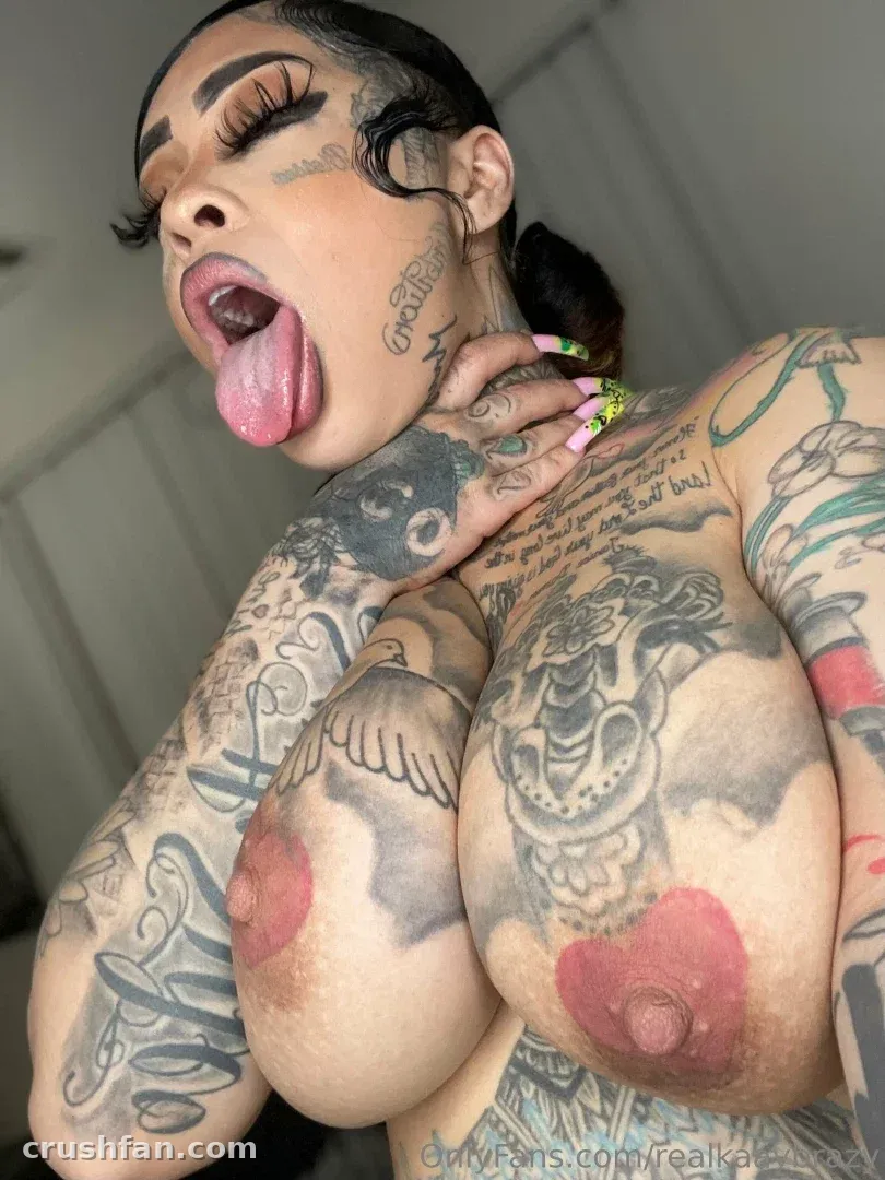 Kaay brazy con la lengua fuera, mostrando tatuajes y uñas de color amarillo verdoso en una selfie de interior.