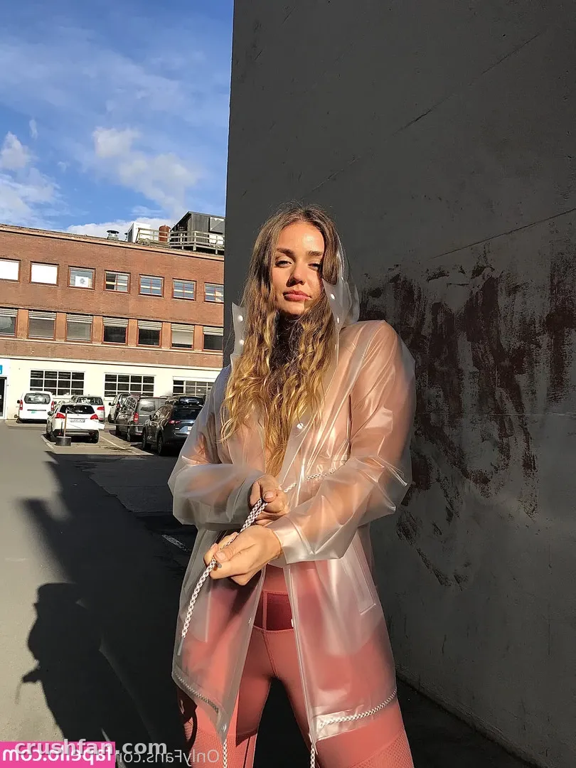 Criadora com capa de chuva transparente e look rosa posando ao lado de uma parede de concreto em uma rua da cidade