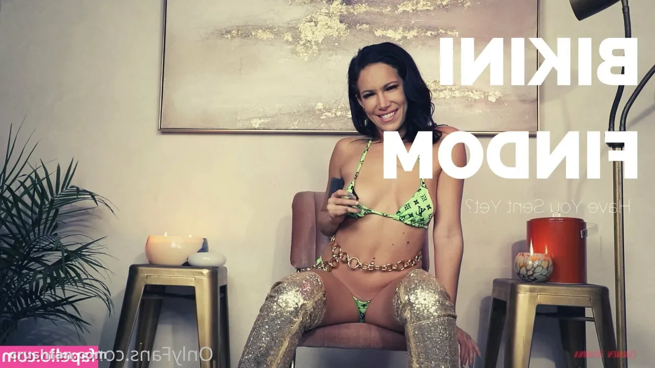Smiling woman in a green bikini and gold leggings seated in a lounge with abstract wall art