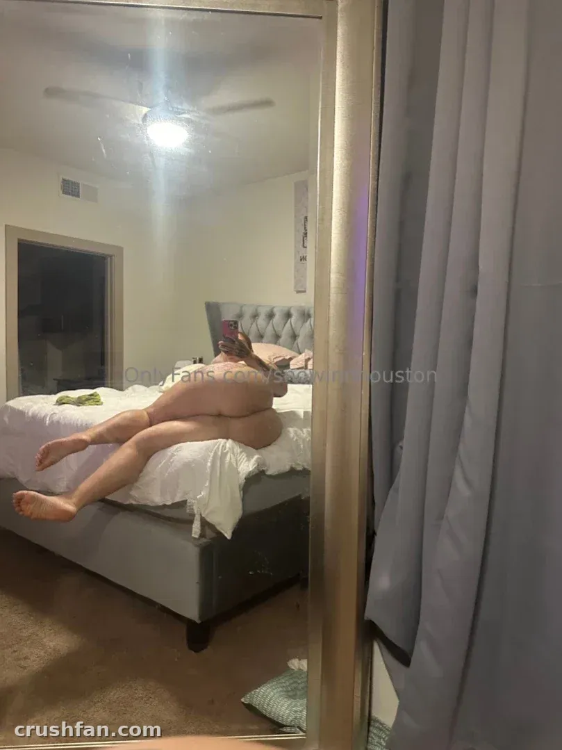 Mirror reflection of Kenzie reclining on a bed in a softly lit bedroom with grey curtains