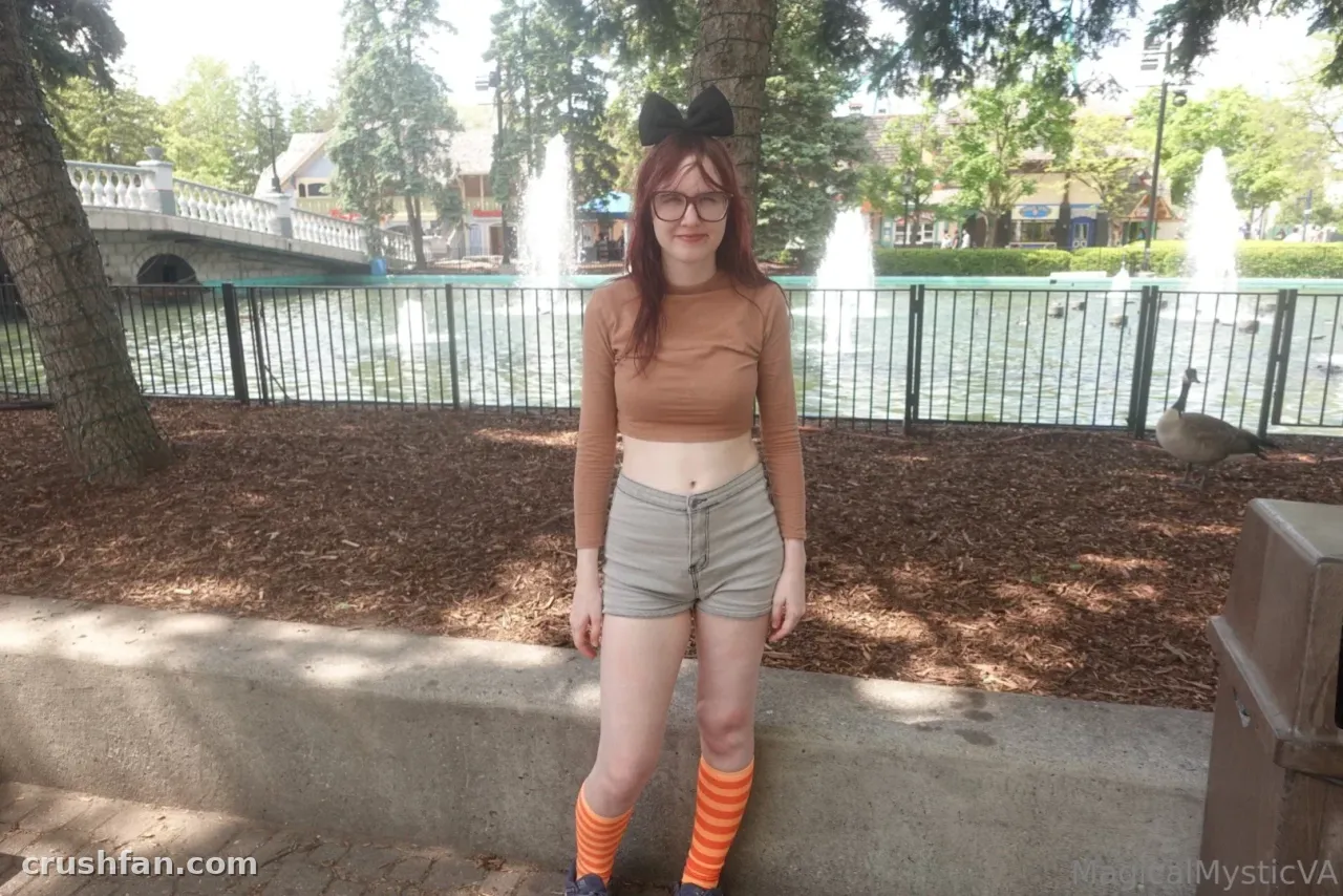 Criadora com top cropped bege e shorts jeans em pé ao lado de um lago de parque com fontes