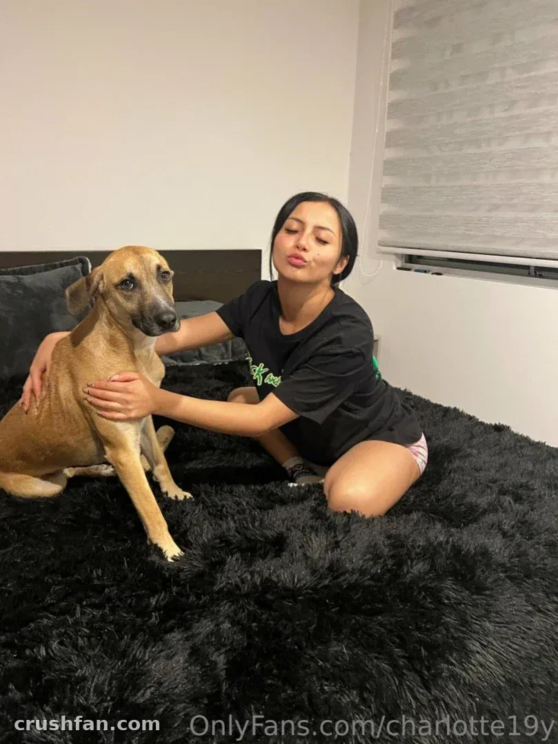 Charlotte in a black t-shirt sitting on a fluffy black bed beside a tan dog, indoors.