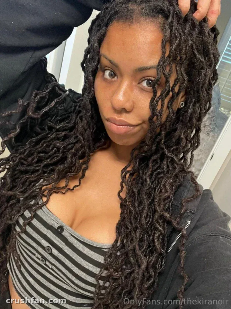 Kira Noir in a striped top and black jacket, looking at the camera with long dark locs.
