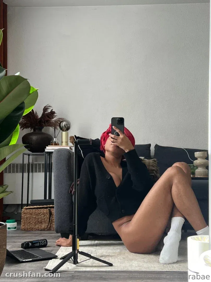 Ts Lira in a black cardigan and white socks sitting on a rug while taking a selfie