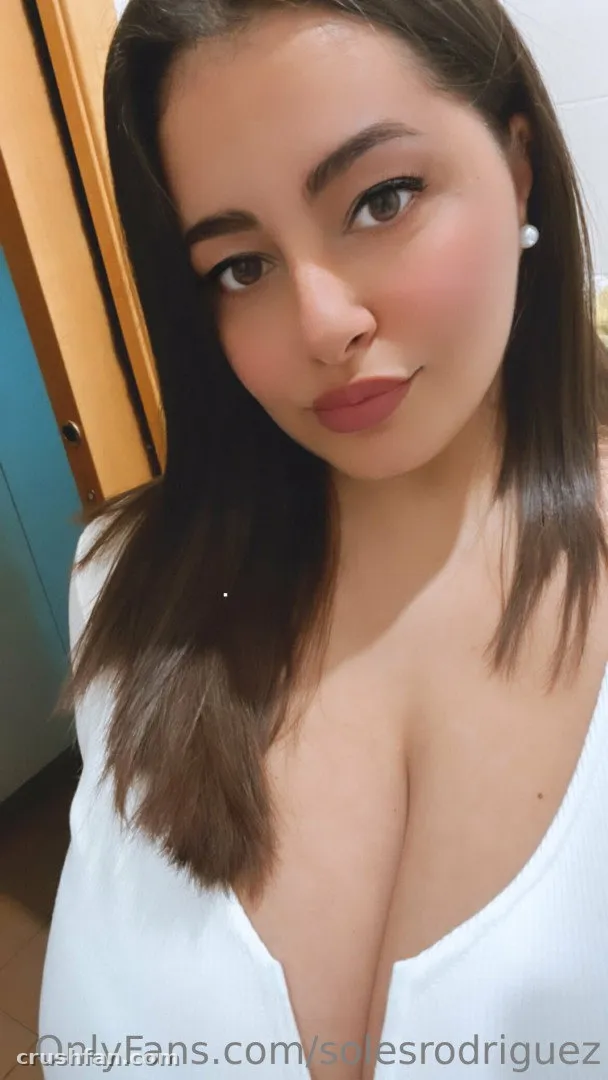 Selfie interna próxima de uma mulher de blusa branca, cabelo longo escuro e iluminação quente