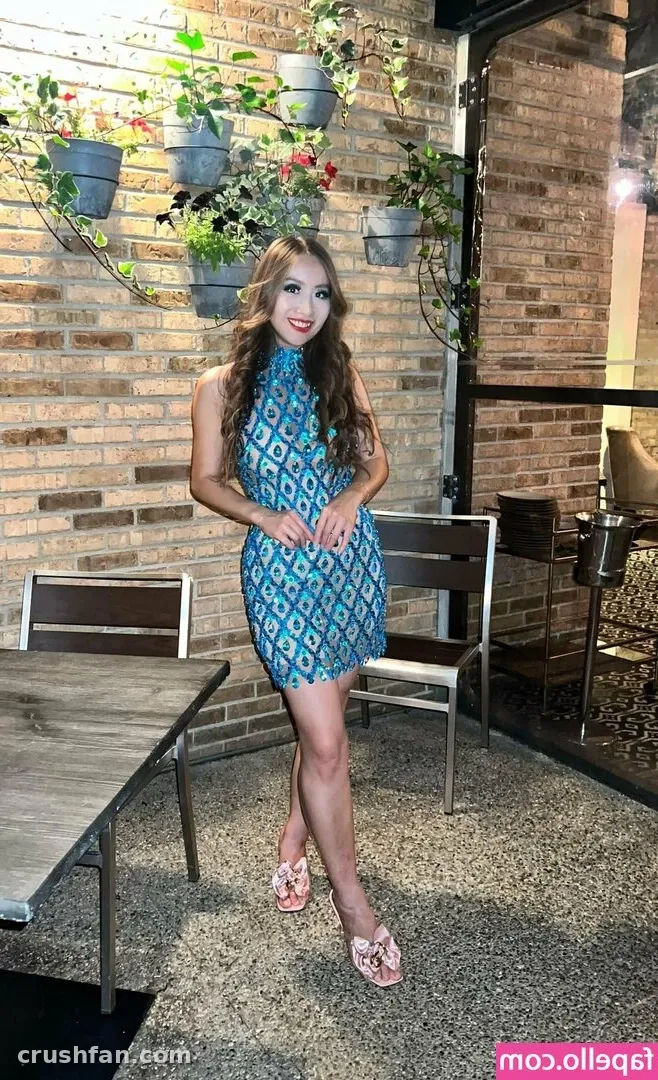 Creator in a blue patterned dress standing on a patio beside wooden tables and a brick wall with plants.