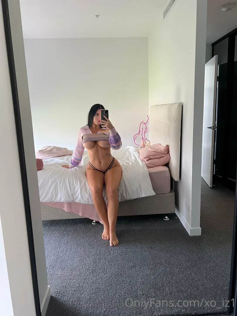 Izzy takes a mirror selfie in a bright bedroom wearing a sheer top and bikini bottoms.
