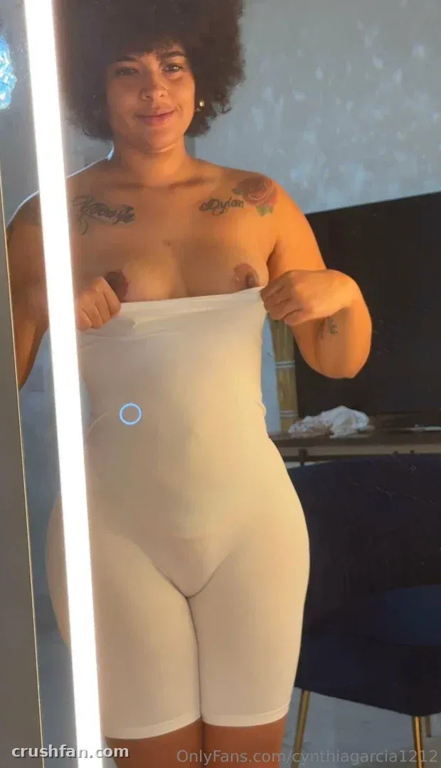 Cynthiagarcia1212 in einem weißen Shaping-Bodysuit vor einem Spiegel stehend.