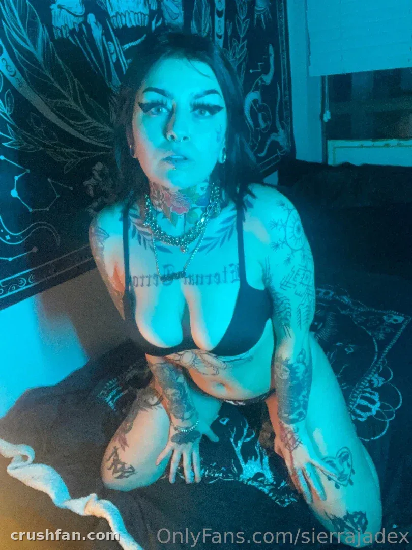 Sierra Jade ajoelhada em uma cama sob luz azul, usando um sutiã preto
