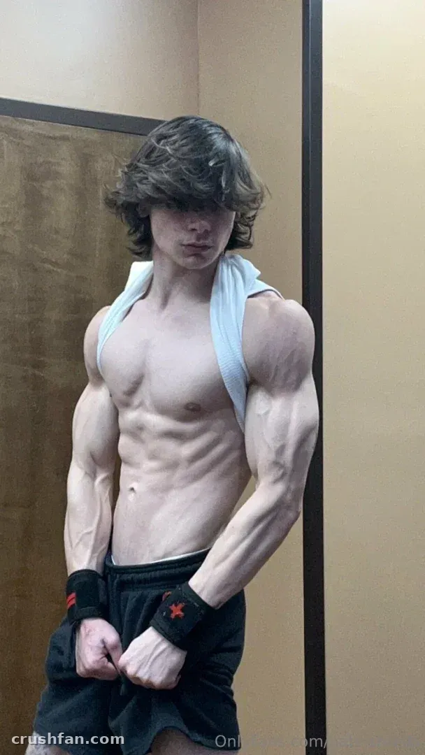 Nathan stands indoors with a light blue towel over his shoulders and dark shorts, flexing his arms.