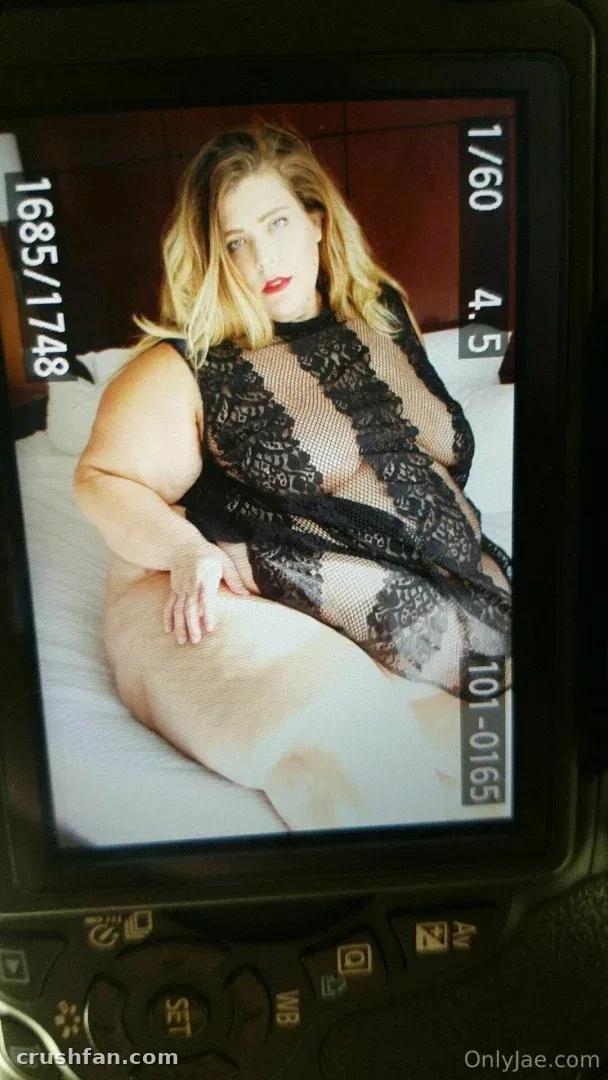 Jae Moonbeam in black lace lingerie seated on a bed, viewed through a camera screen.