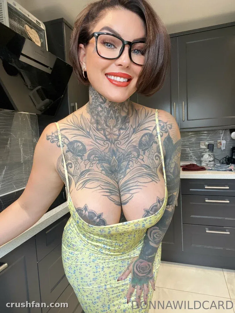Creator in einem gelben Blumenkleid und Brille, stehend in einer Küche mit sichtbaren Tattoos.