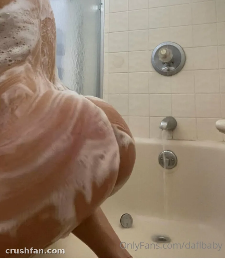 Da FL Baby leaning in a bathtub filled with white bubbles, with beige tiles and silver fixtures behind.