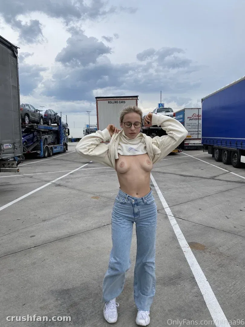 Nina Joy in Jeans und weißen Sneakern auf einem Betonplatz mit Lastwagen hinter sich