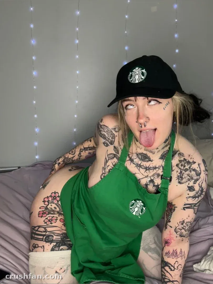 Furbee con delantal verde y gorra negra sobre una cama, lengua fuera, mostrando tatuajes