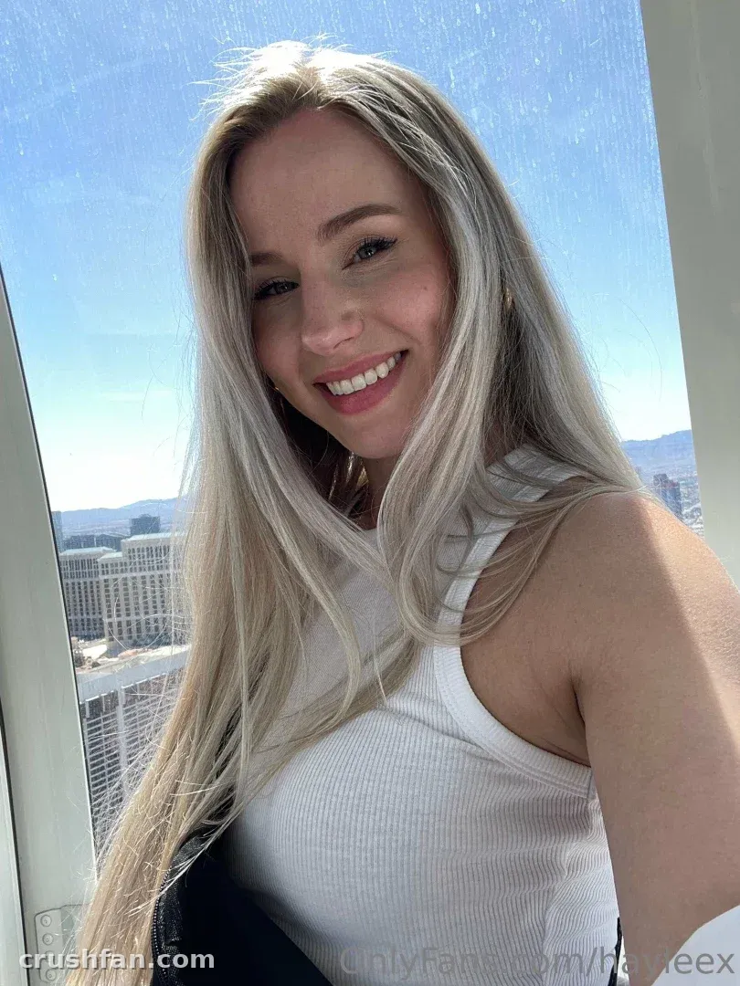 Hayleex smiling in a white tank top during a bright selfie by a window with a city view.