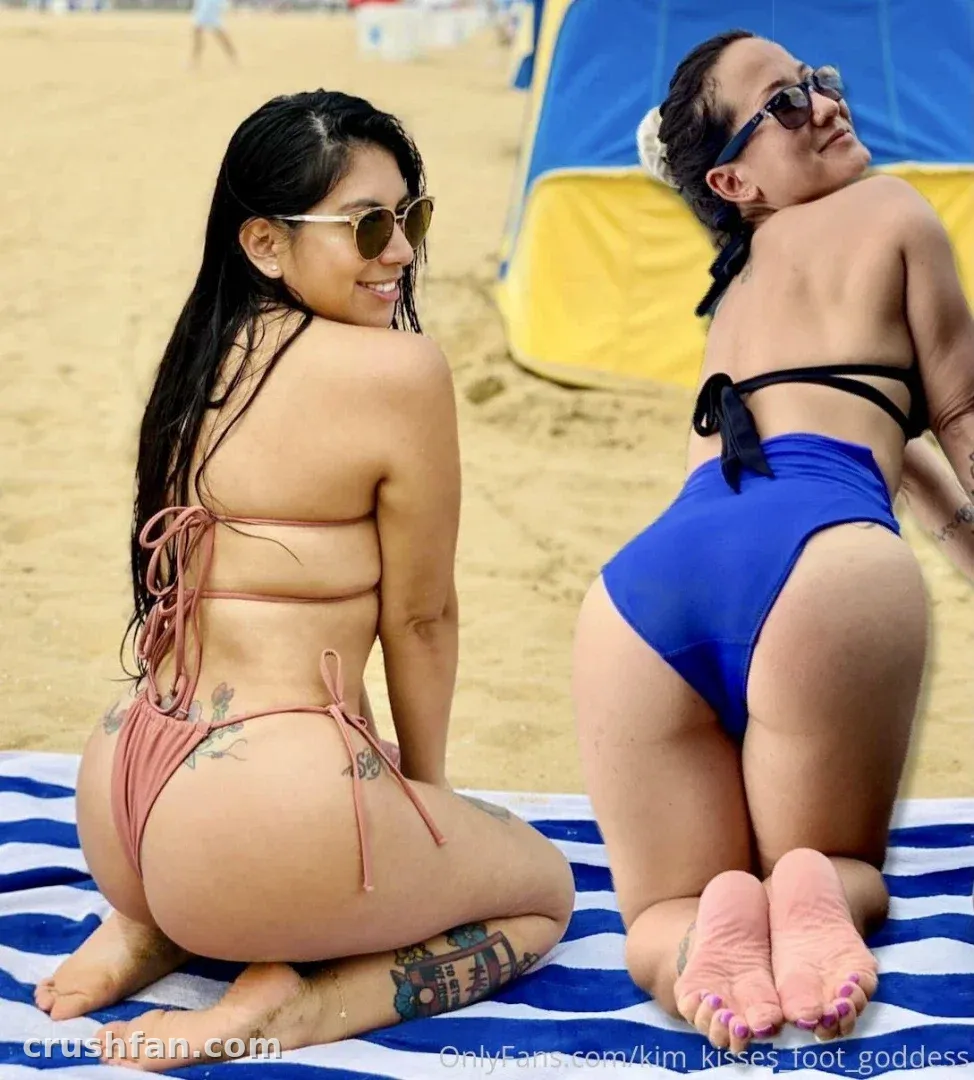 Kim Kisses kneeling on a striped beach towel in a pink bikini under bright sunlight