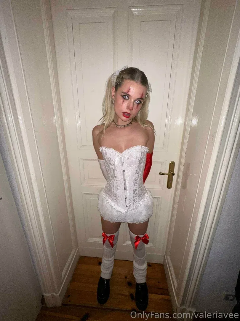 Mona Marie com um vestido de corset branco e meias em pé em um corredor com portas brancas.