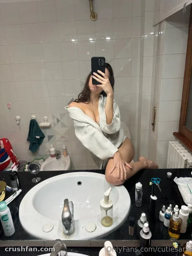 CutieSara prenant un selfie miroir dans une salle de bain portant un pull blanc à épaules dénudées.