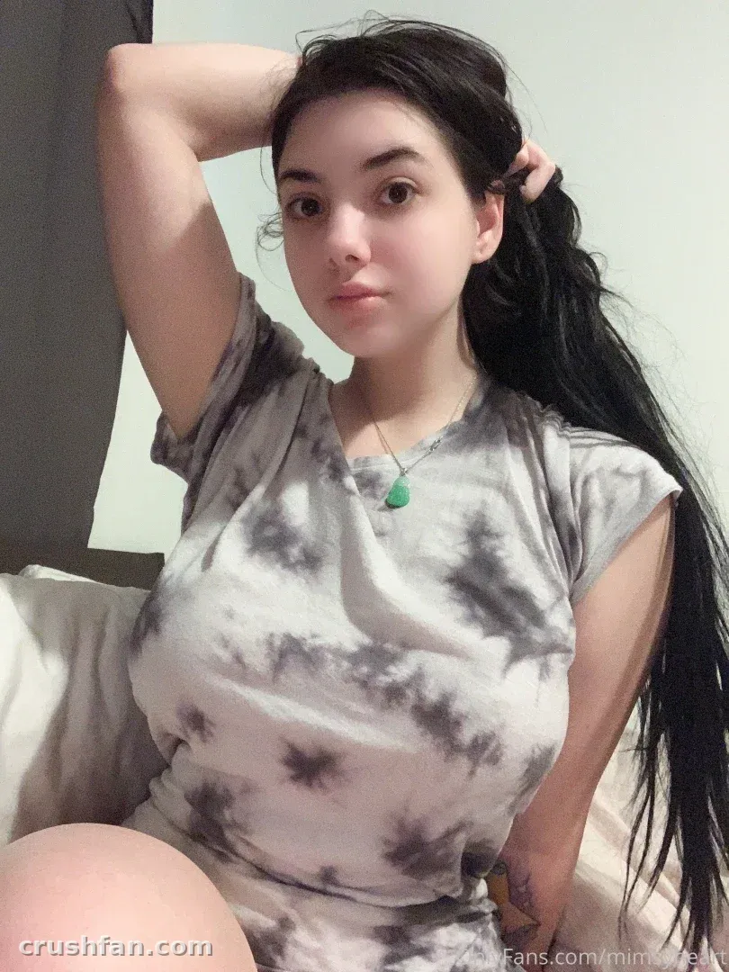 Amber com um top tie-dye cinza e um pingente verde, sentada em ambiente interno contra uma parede clara.