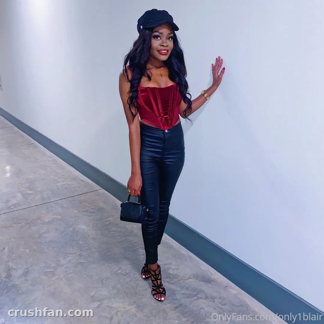 Blair com um corset de veludo vermelho e calça de couro preta posando em um corredor claro