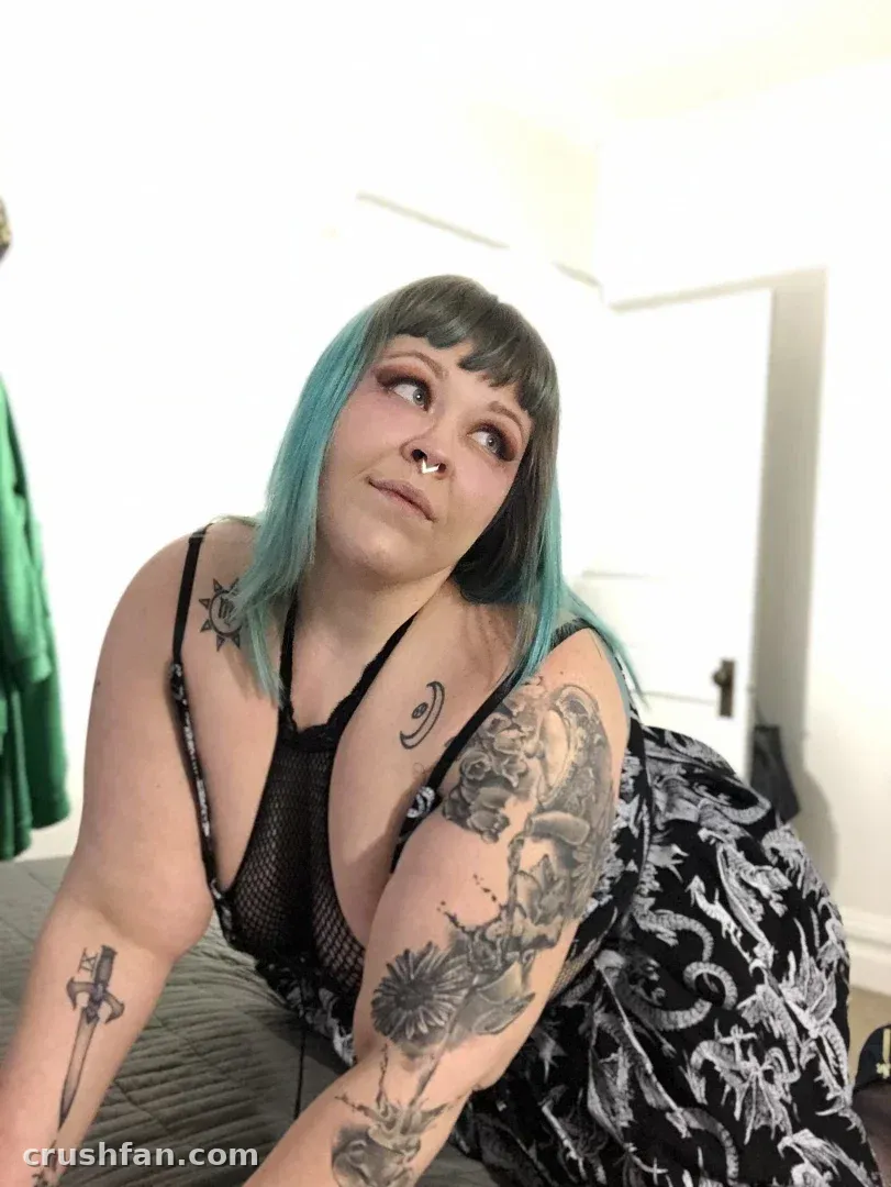 Creator with teal-tipped hair and tattoos in a black mesh top, looking upward indoors.