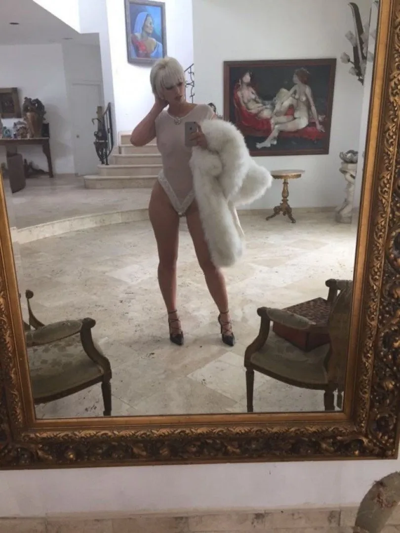 Yesjulz takes a mirror selfie in a light bodysuit and white fur inside a bright room.