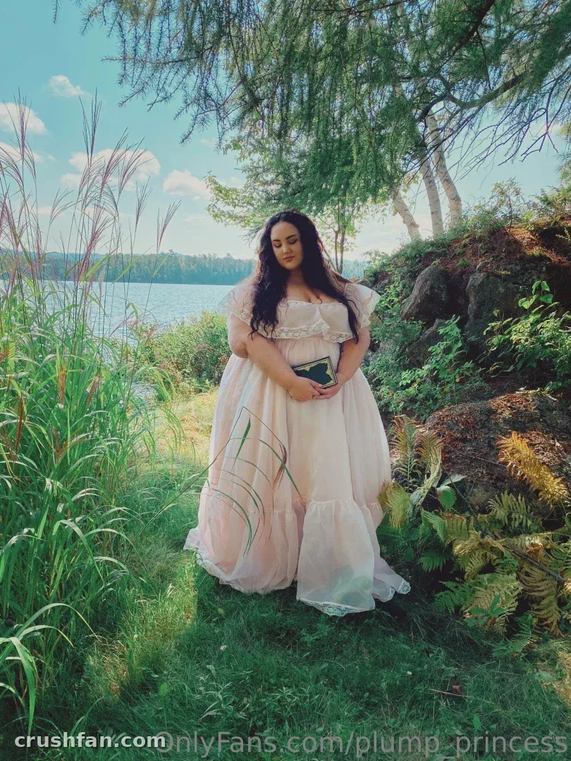 Plump Princess in a blush gown standing by a lake among tall grasses and trees