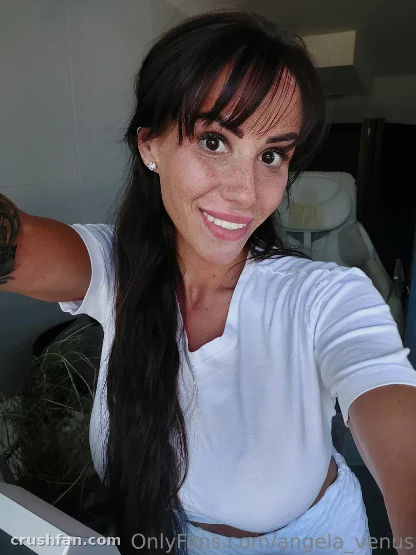 Angela souriant avec un haut blanc lors d'un selfie en intérieur