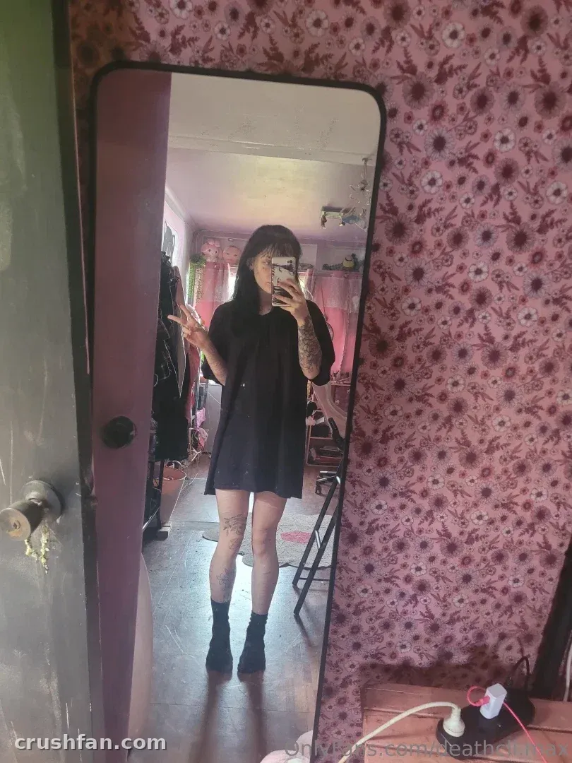 Maxine con un vestido negro tomándose una selfie frente al espejo en una habitación con papel pintado floral rosa.