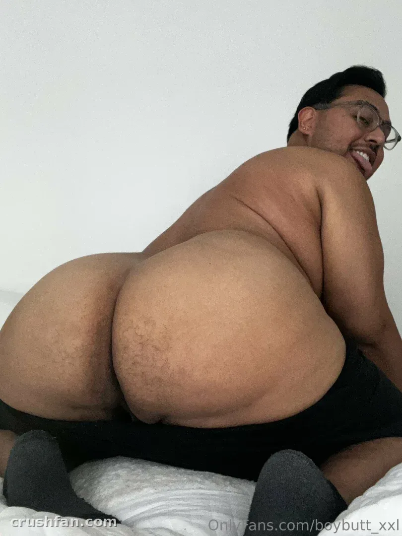 BoyButtXXL arrodillado en una cama blanca, mirando hacia atrás con una sonrisa