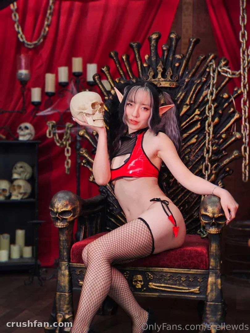 Creator in rotem Latex und Netzstrümpfen auf einem schädelverzierten Thron sitzend, einen Schädel als Requisite haltend