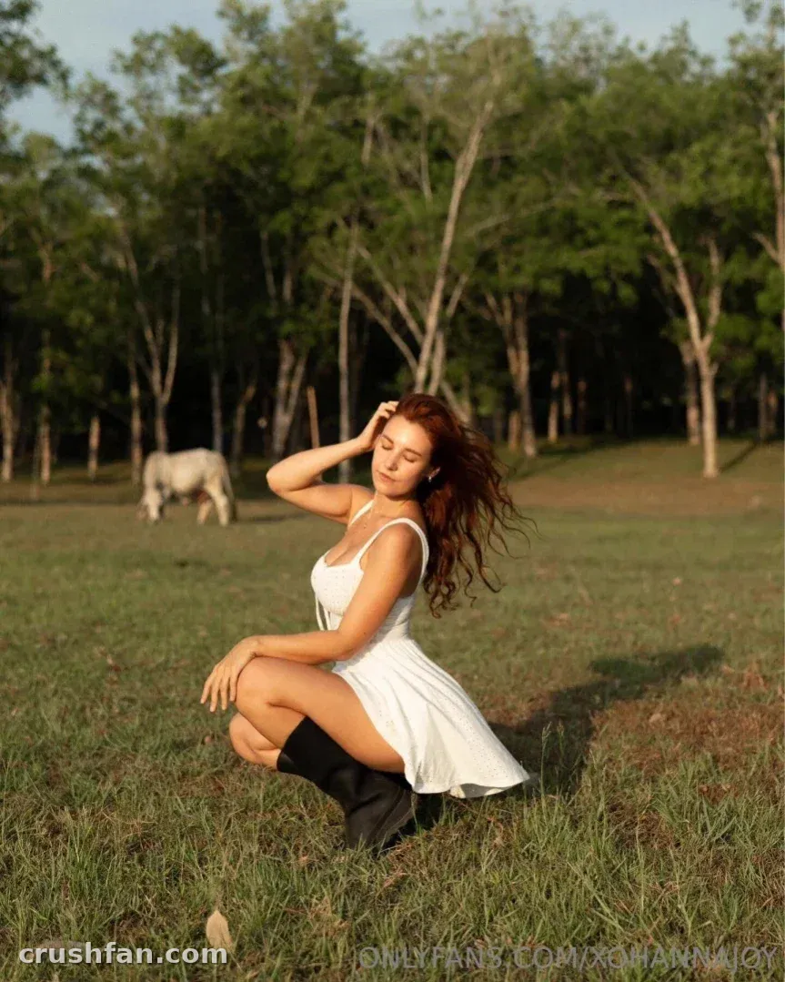 Creator en robe blanche et bottes noires, accroupie dans une prairie ensoleillée avec des arbres et un cheval derrière elle.