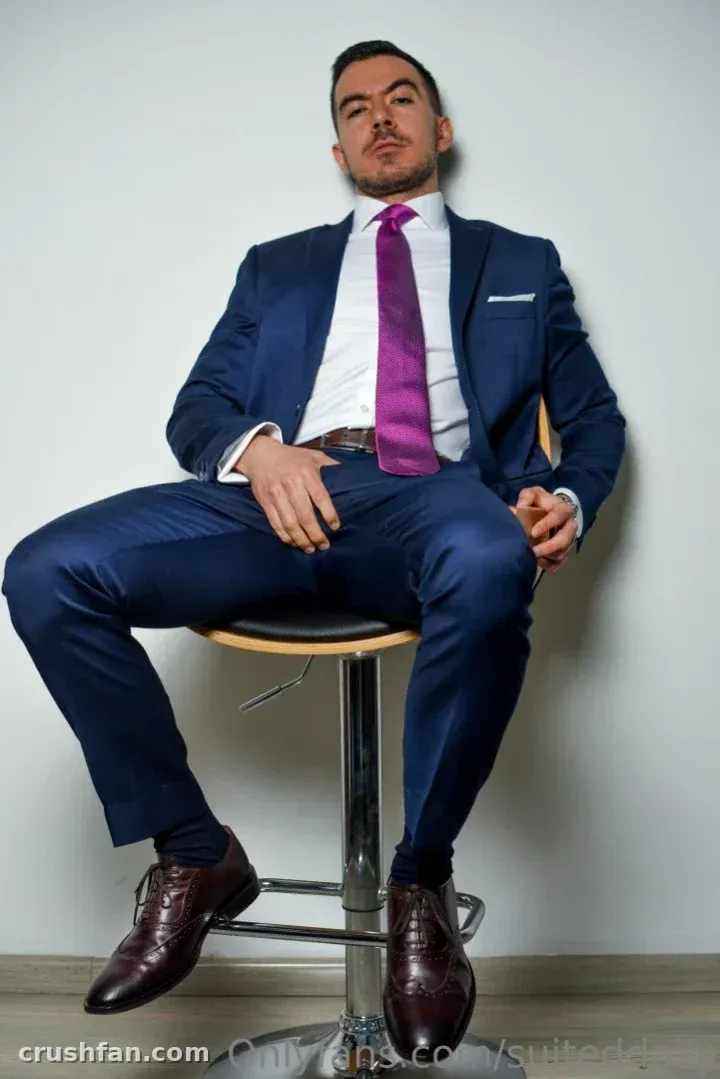 Man in a navy suit and purple tie seated on a chrome bar stool against a plain wall