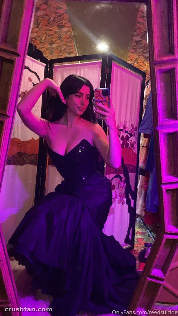 Reed in a dark strapless gown taking a mirror selfie in purple light