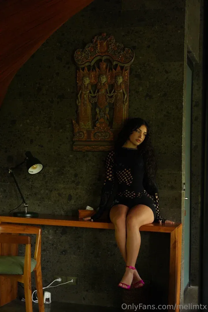 Melissa com um conjunto preto de bolinhas e saltos rosa sentada sobre uma mesa de madeira em um quarto com pouca luz