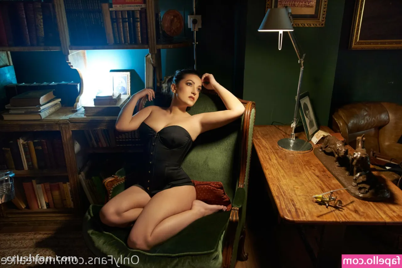 Alice in a black corset posing in a vintage study with bookshelves and a desk lamp