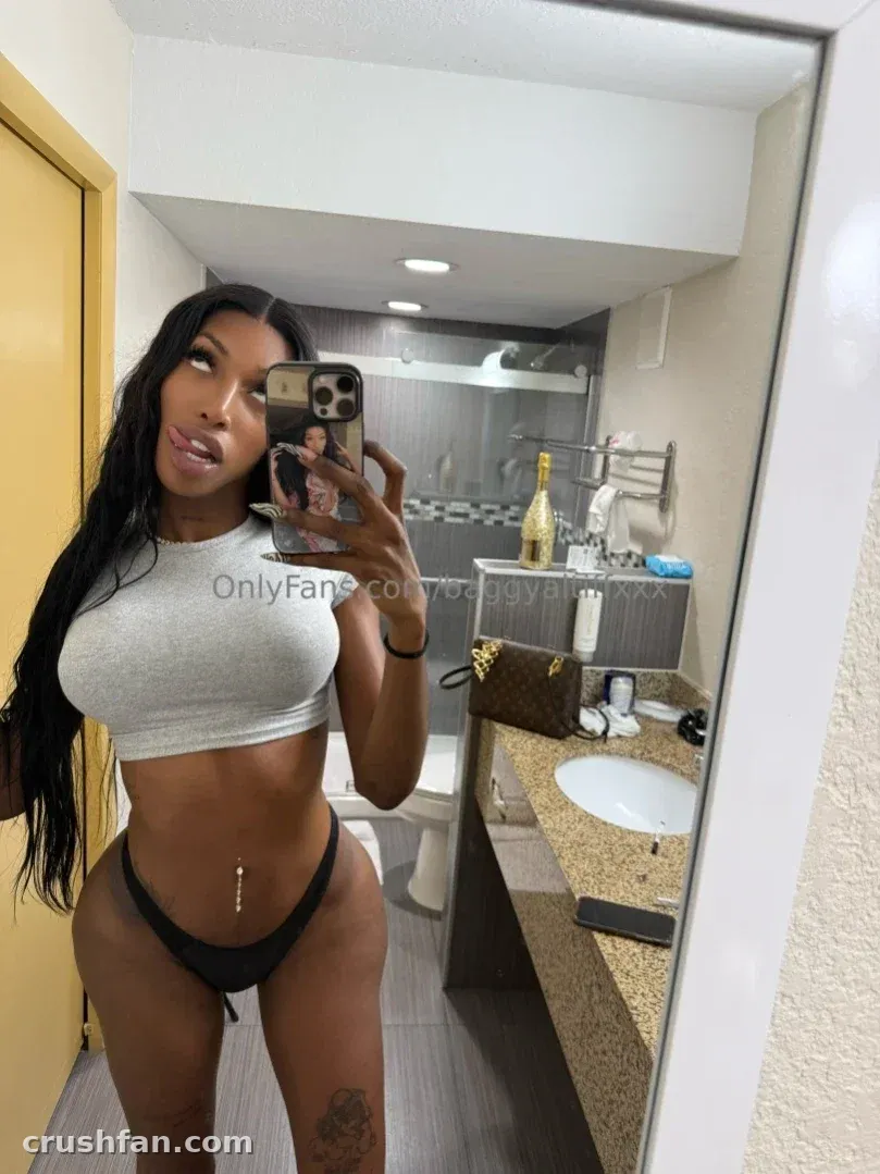 mAmi-Tiff tirando uma selfie no espelho de um banheiro usando um top cropped cinza e uma peça inferior preta.