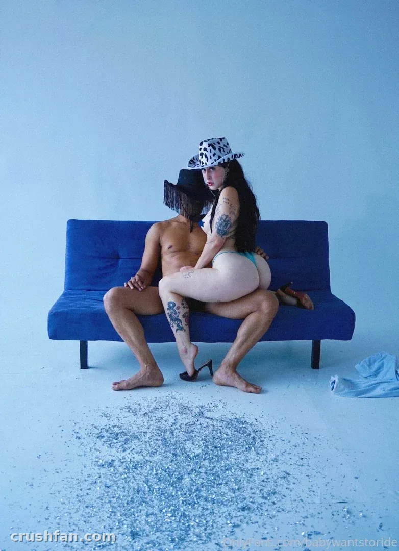 Créatrice avec un chapeau à motifs assise sur un canapé bleu dans un studio lumineux avec des fragments dispersés sur le sol.