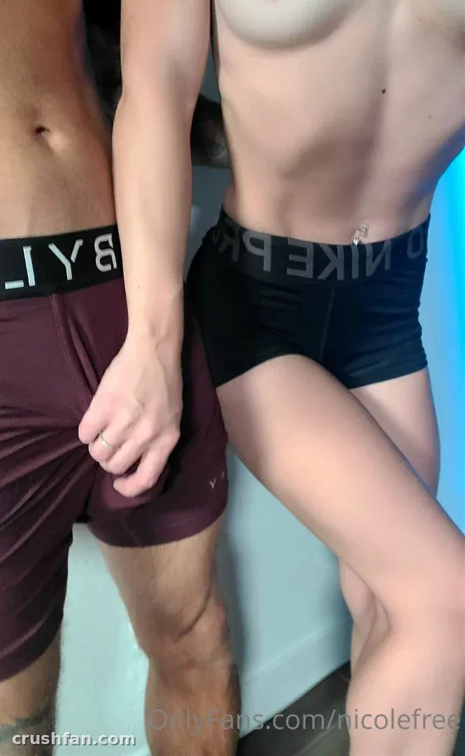 Nicole FREE stands beside another person in dark boxer briefs and maroon shorts against a blue wall.