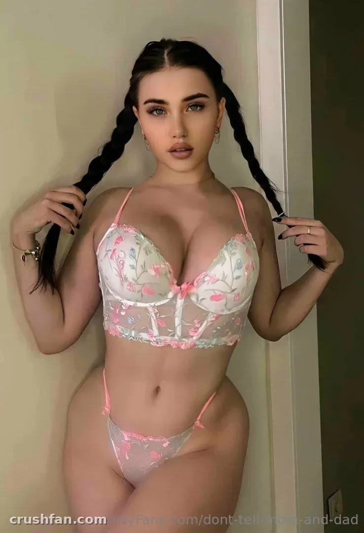 Creator in white floral lingerie holding two braids, standing against a plain wall