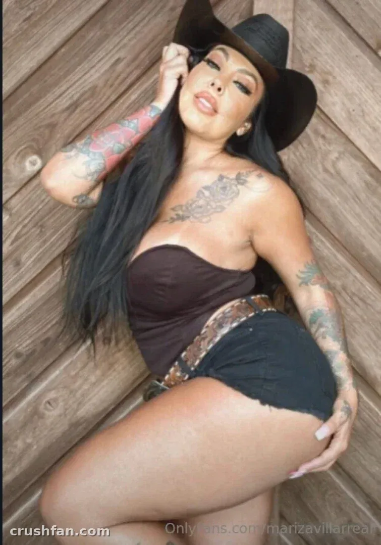 Mariza Villarreal com um chapéu preto e top escuro posando ao lado de uma parede de madeira