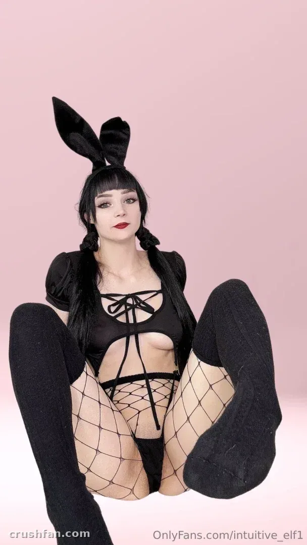 That Elf Lady com uma roupa de coelhinha preta e meia-calça de rede contra um fundo rosa