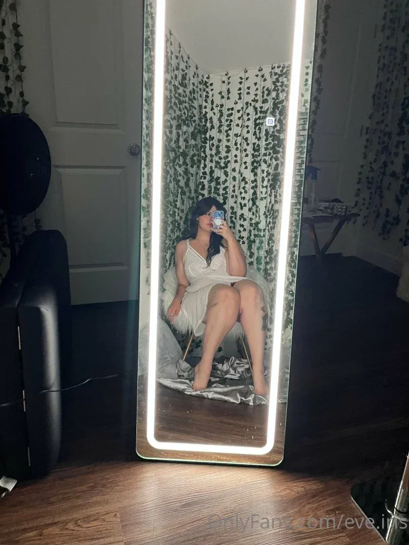 Eve Iris dans une robe nuisette blanche prenant un selfie miroir dans une pièce avec un papier peint à feuilles.