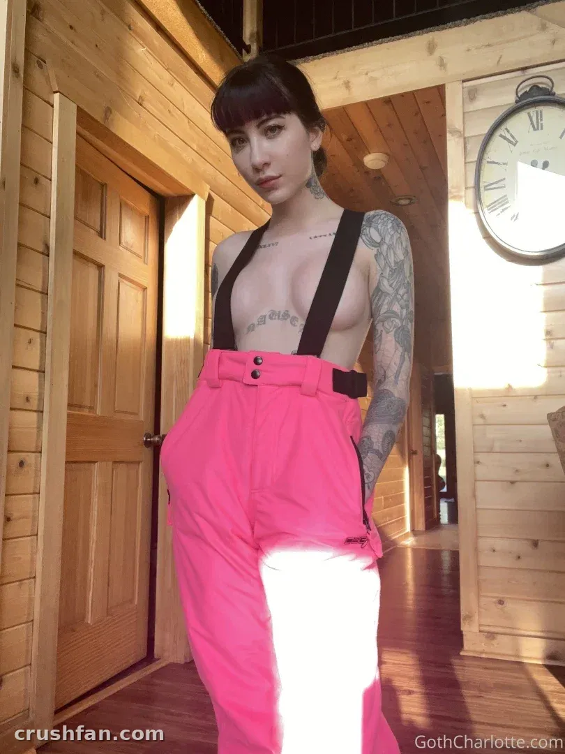 Goth Charlotte en pantalon rose à taille haute et bretelles dans une pièce en bois.