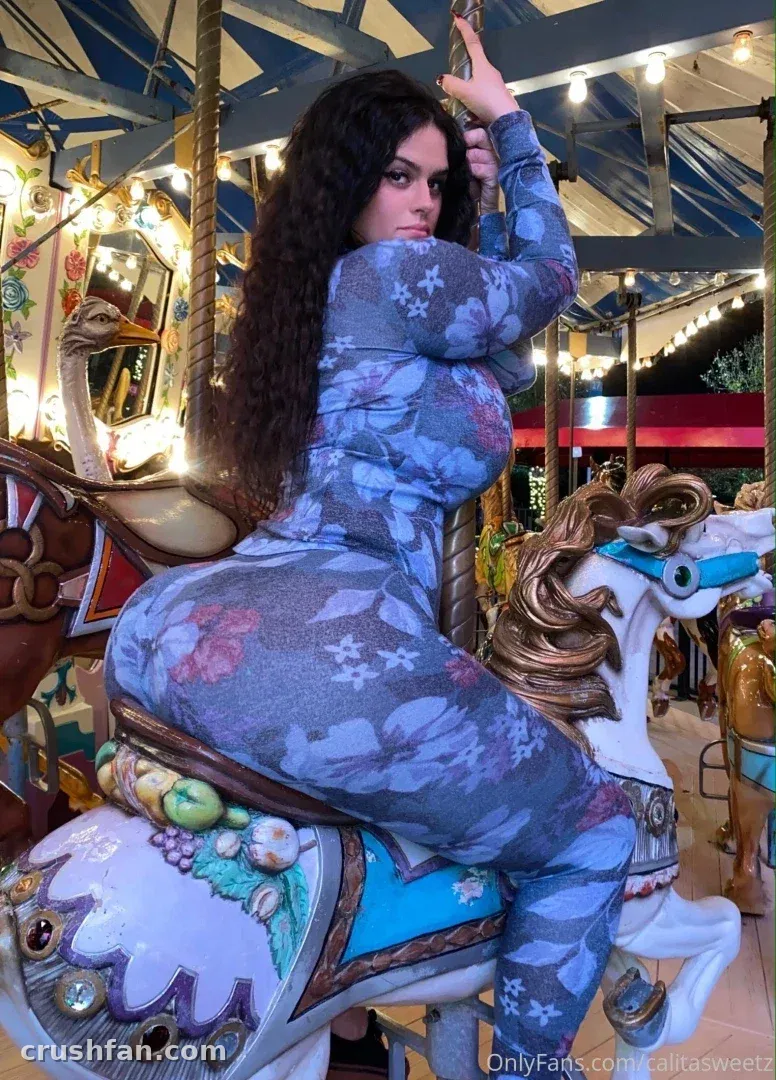 Bootyy Princess em um catsuit floral sentada em um cavalo de carrossel sob luzes quentes.