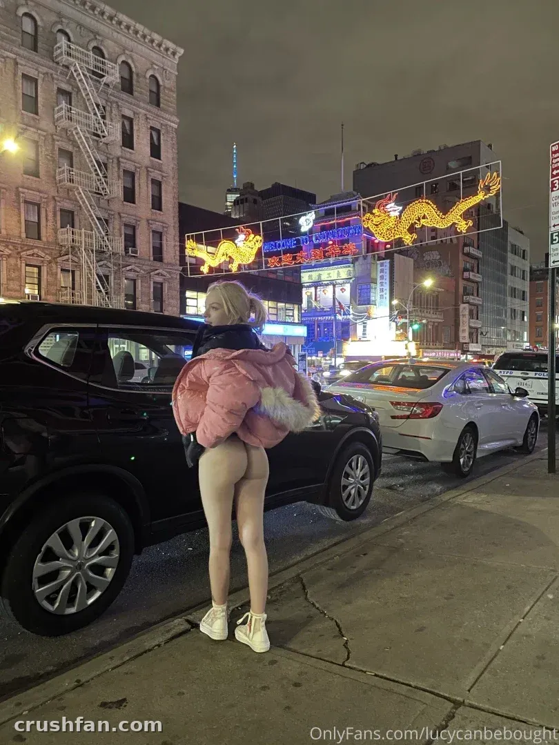 Lucy in a pink puffer jacket standing on a night city sidewalk beside a parked car