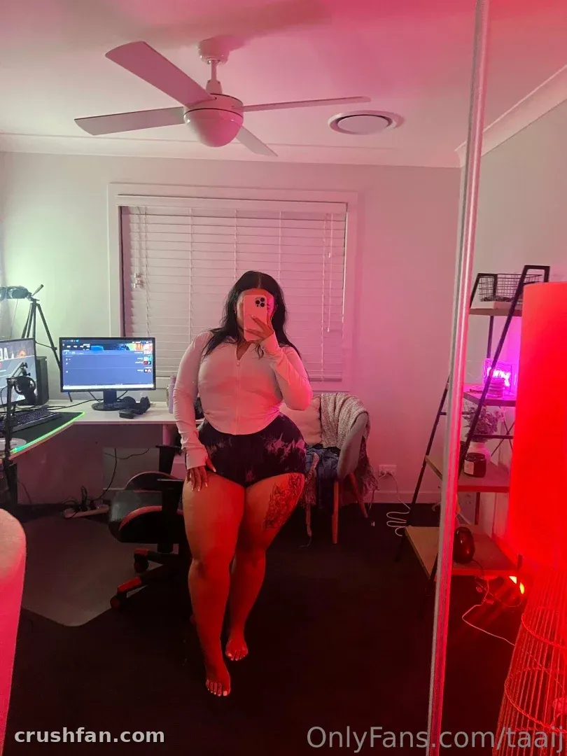 TAAIJ takes a mirror selfie in a pink-lit room with a desk and ceiling fan behind her.
